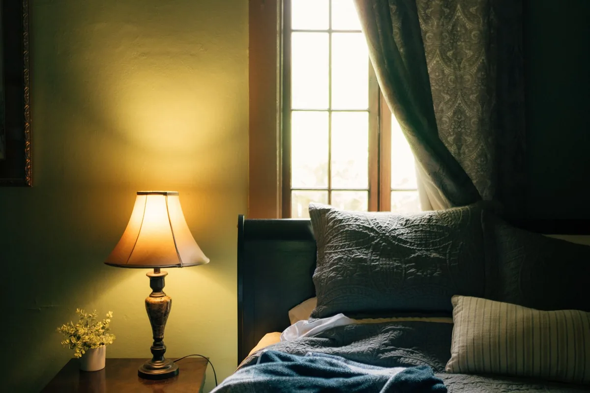 A lit table lamp and a small potted plant on a nightstand beside an unmade bed with grey pillows, by a tall window with a patterned curtain