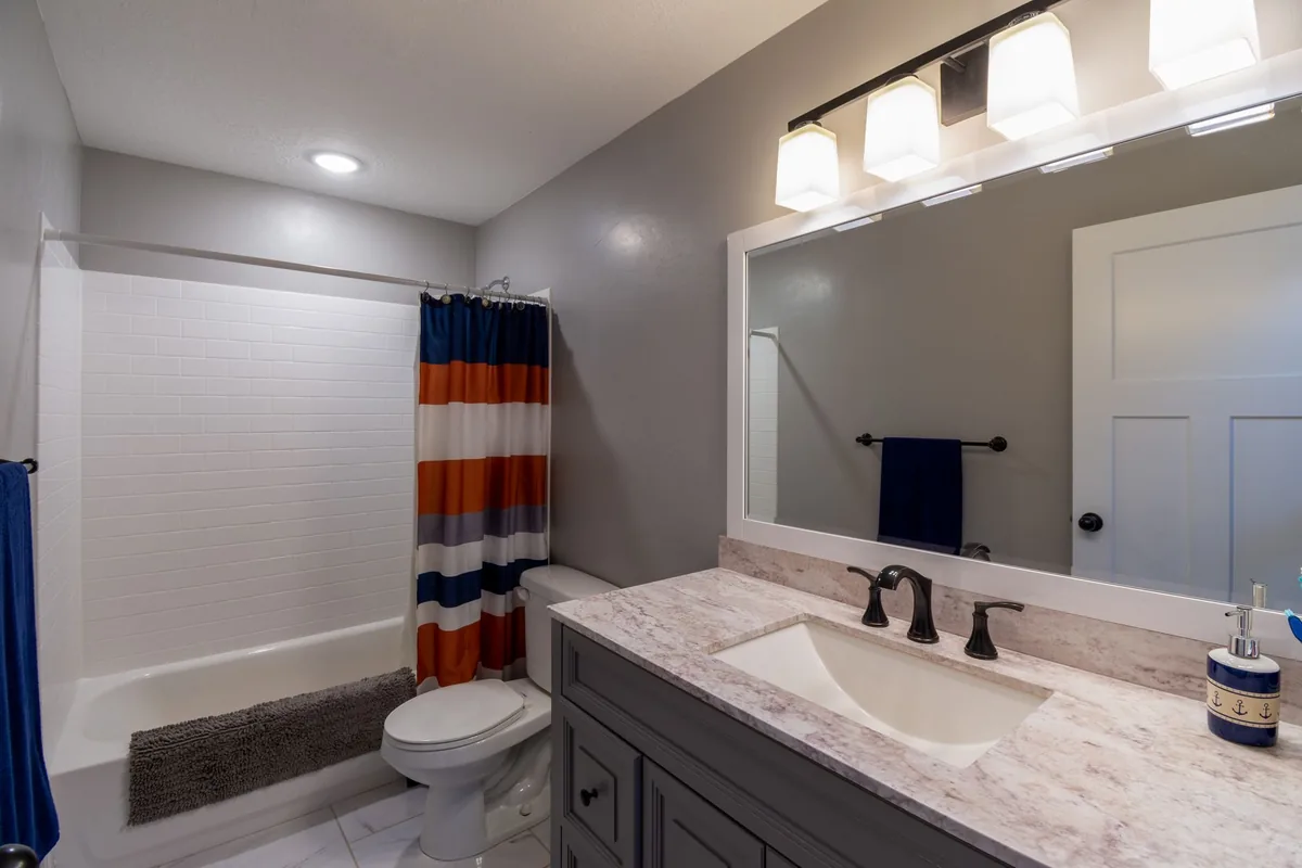 A small bathroom with a white tub behind a striped shower curtain, a toilet and a grey vanity with a wide mirror