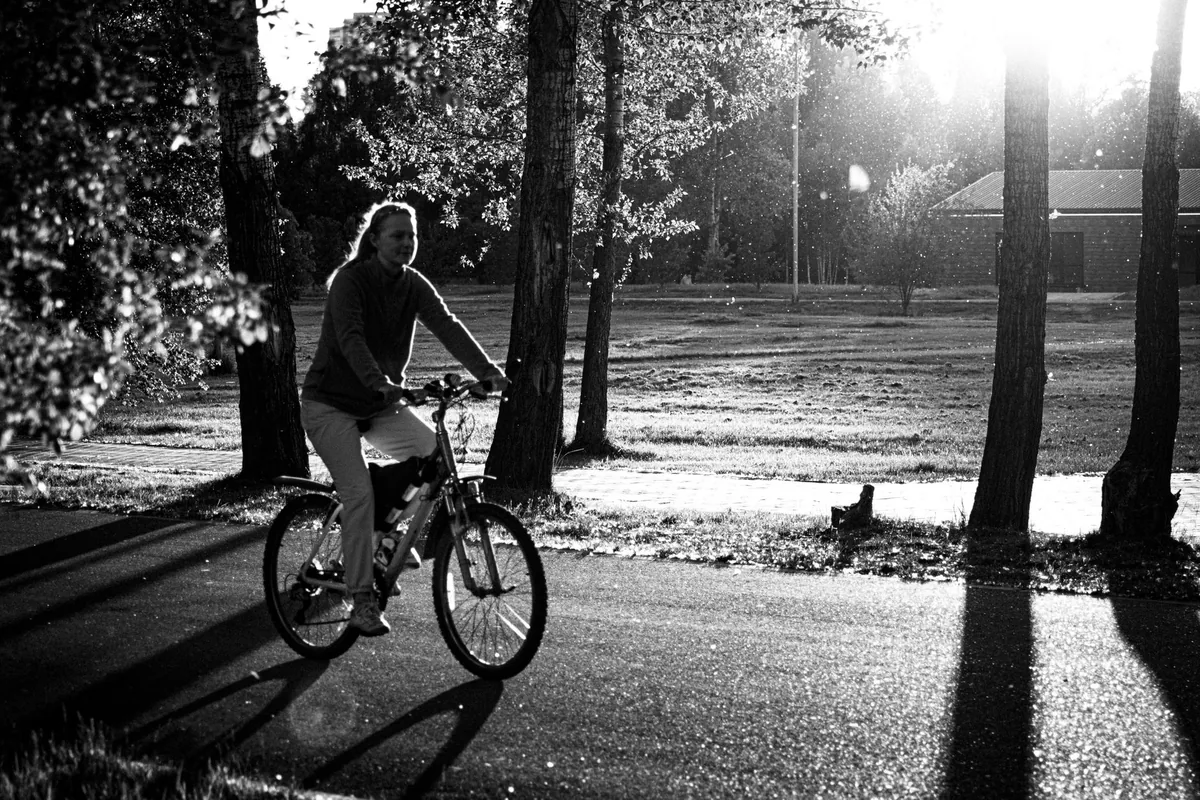 A woman riding a bicycle along a paved path through a sunlit park, in black and white