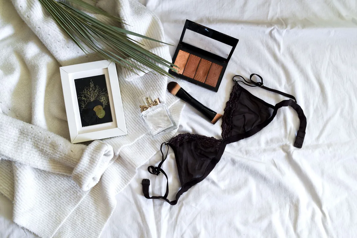 A black lace bra with its hook-and-eye fastening laid on a white bed next to a makeup palette, a brush and a small framed print