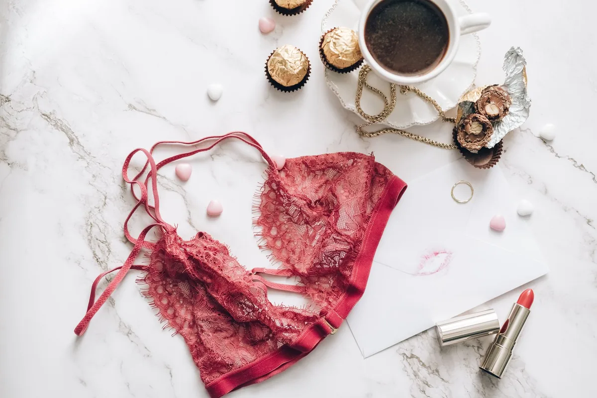 A red lace bralette laid flat on a white marble surface next to a coffee cup, chocolates and a lipstick