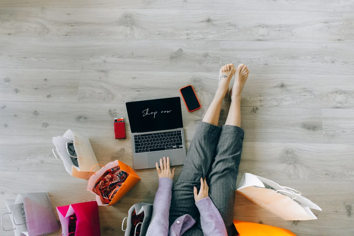 A woman sitting on a wooden floor with a laptop surrounded by paper shopping bags, seen from above