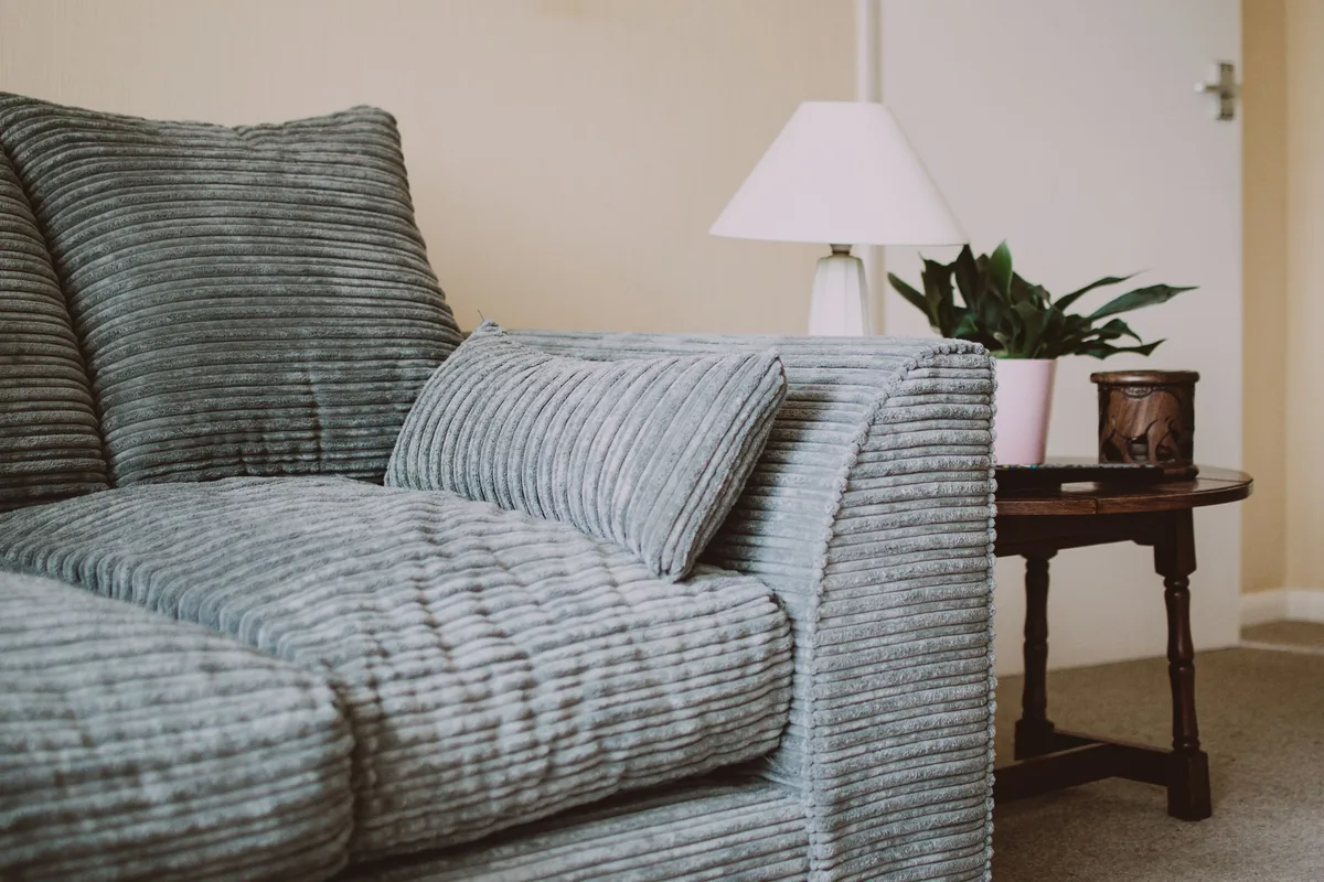 The arm and seat cushion of a grey jumbo cord sofa beside a small wooden side table and a lamp