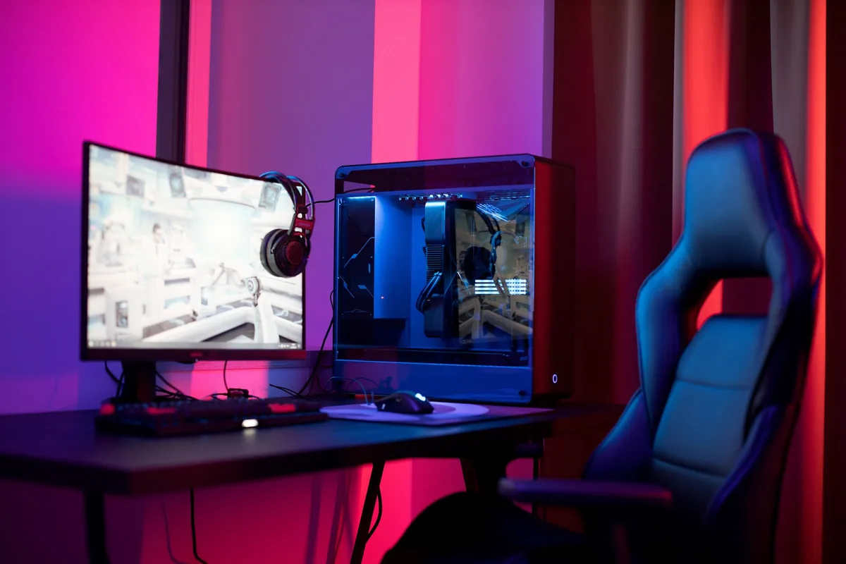 An empty high-backed chair beside a desk with a curved monitor, headphones and a glass-sided computer case, lit pink and blue