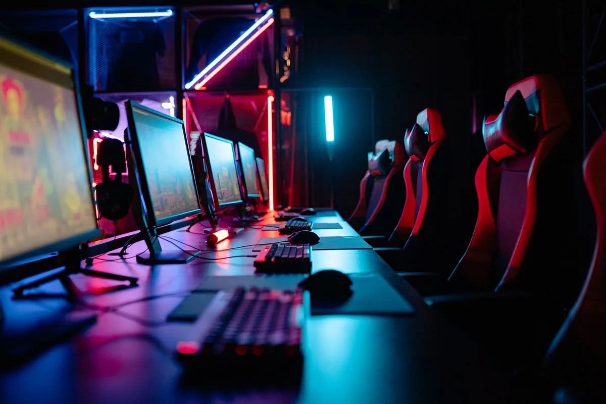 A row of red and black high-backed chairs facing a long desk of monitors and keyboards in a dark room lit by neon strips