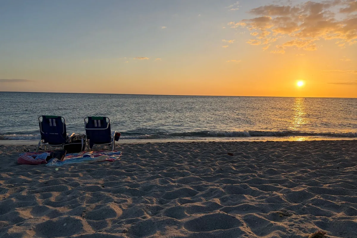 Two empty folding beach chairs on the sand facing the sea at sunset, with a towel and a bag on the ground beside them