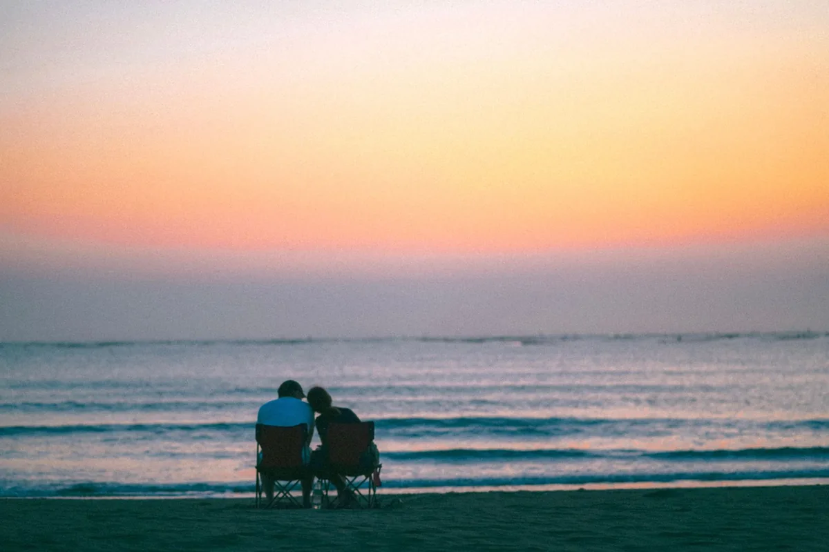Two people seen from behind sitting in folding chairs on a beach at dusk, facing small waves under a pink sky