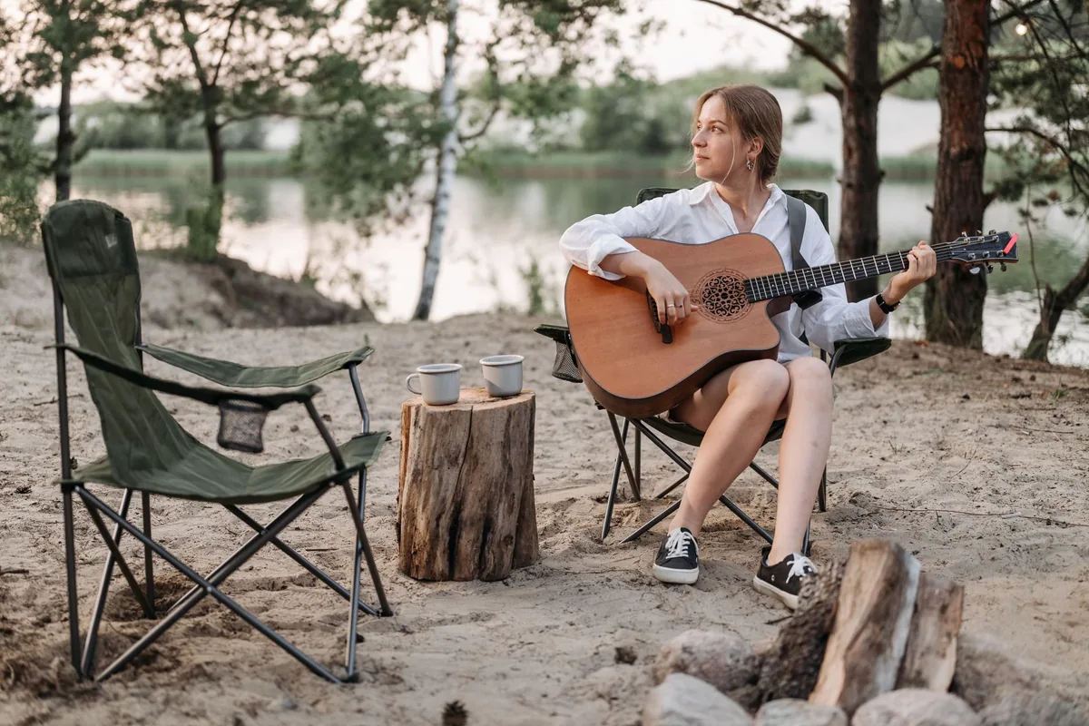 An empty green folding quad chair beside a woman playing guitar in a second chair on a sandy lakeshore