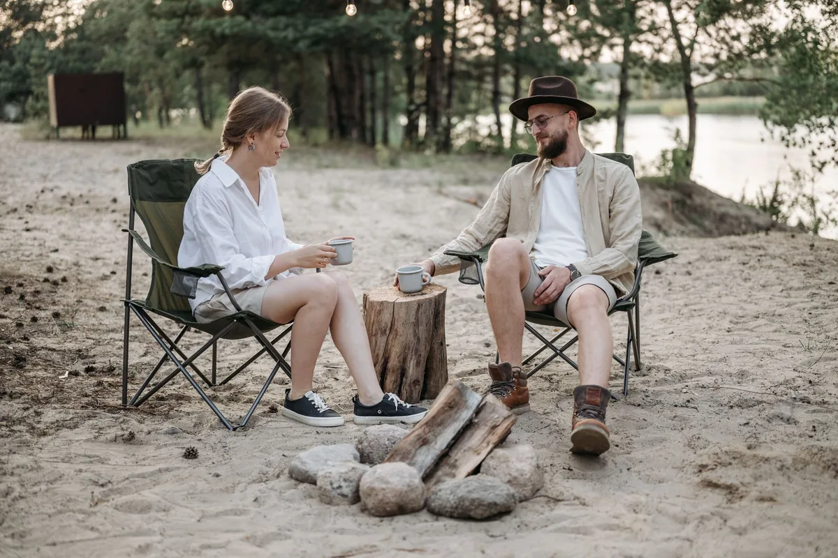 Two people sitting in folding quad chairs beside a stone fire ring on a sandy lakeshore, holding enamel mugs