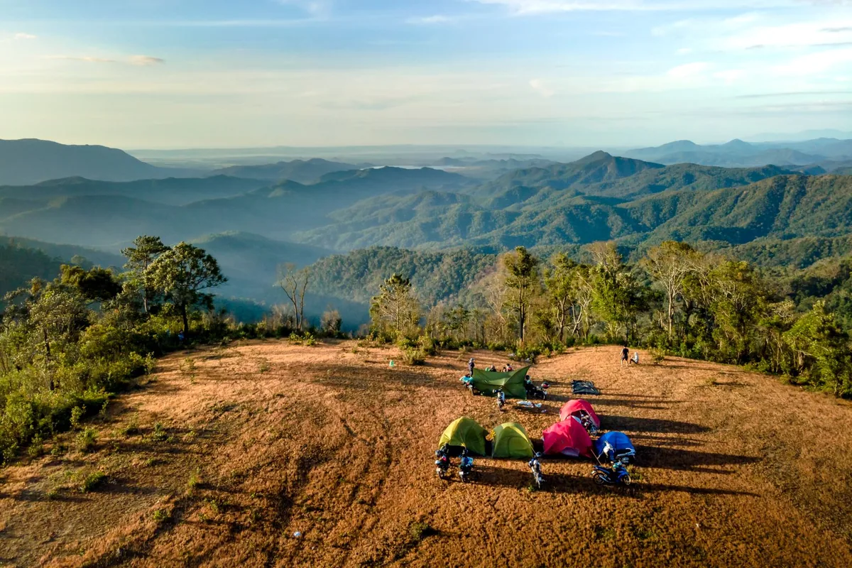 Aerial view of small tents pitched on a cleared hilltop above forested mountains at sunrise