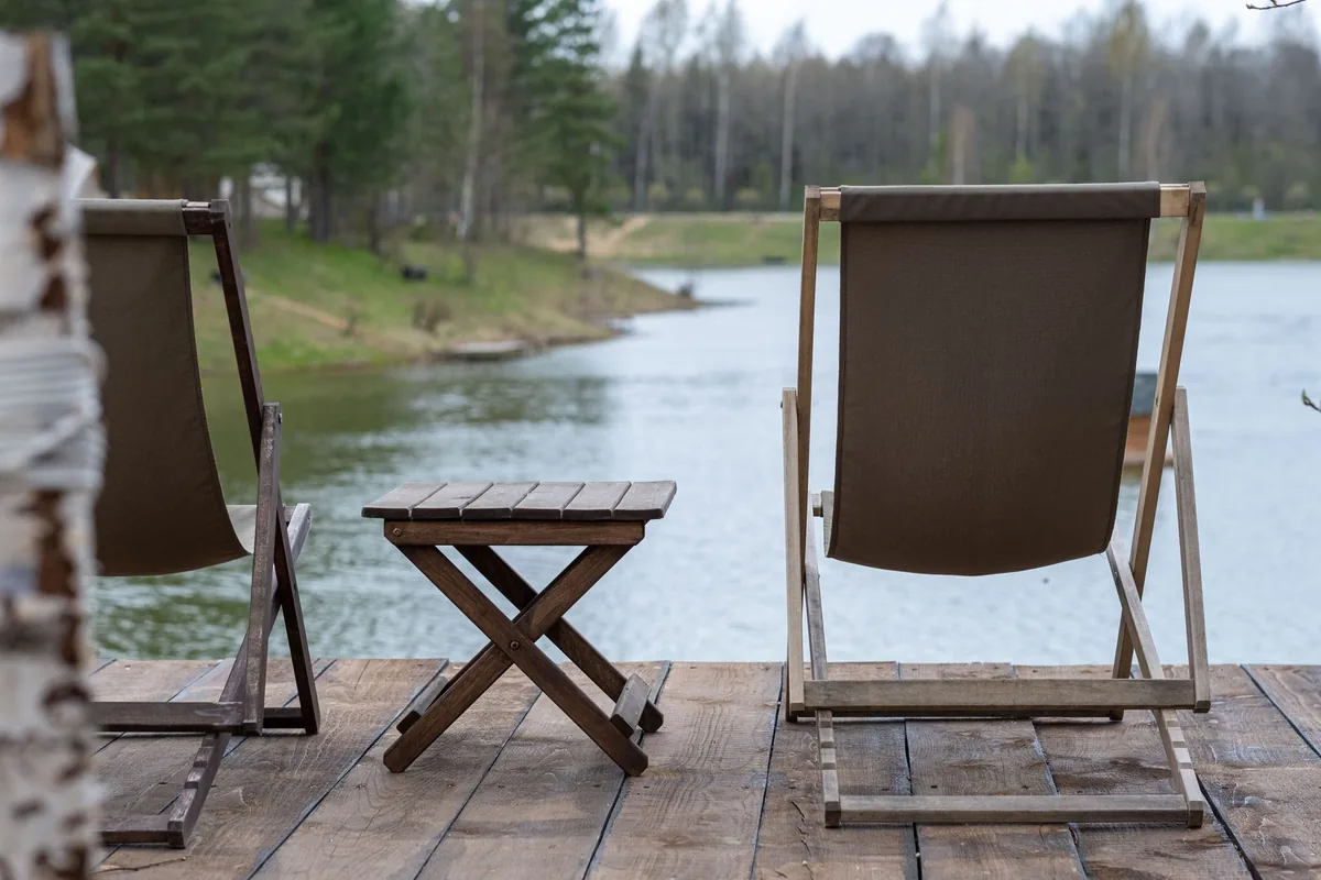 Two folding wooden sling chairs with brown fabric and a small slatted table on a deck by a lake