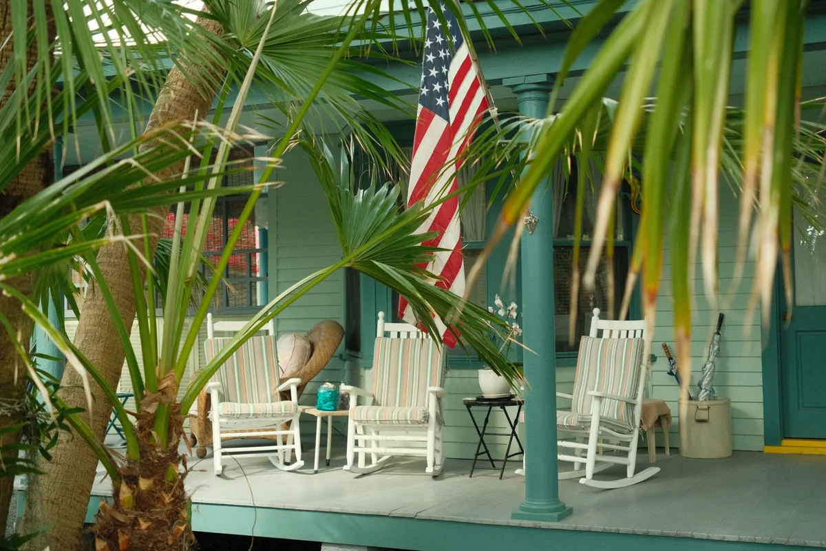 Three white rocking chairs with striped seat and back cushions on a painted porch behind palm fronds, with a flag hanging from a porch post
