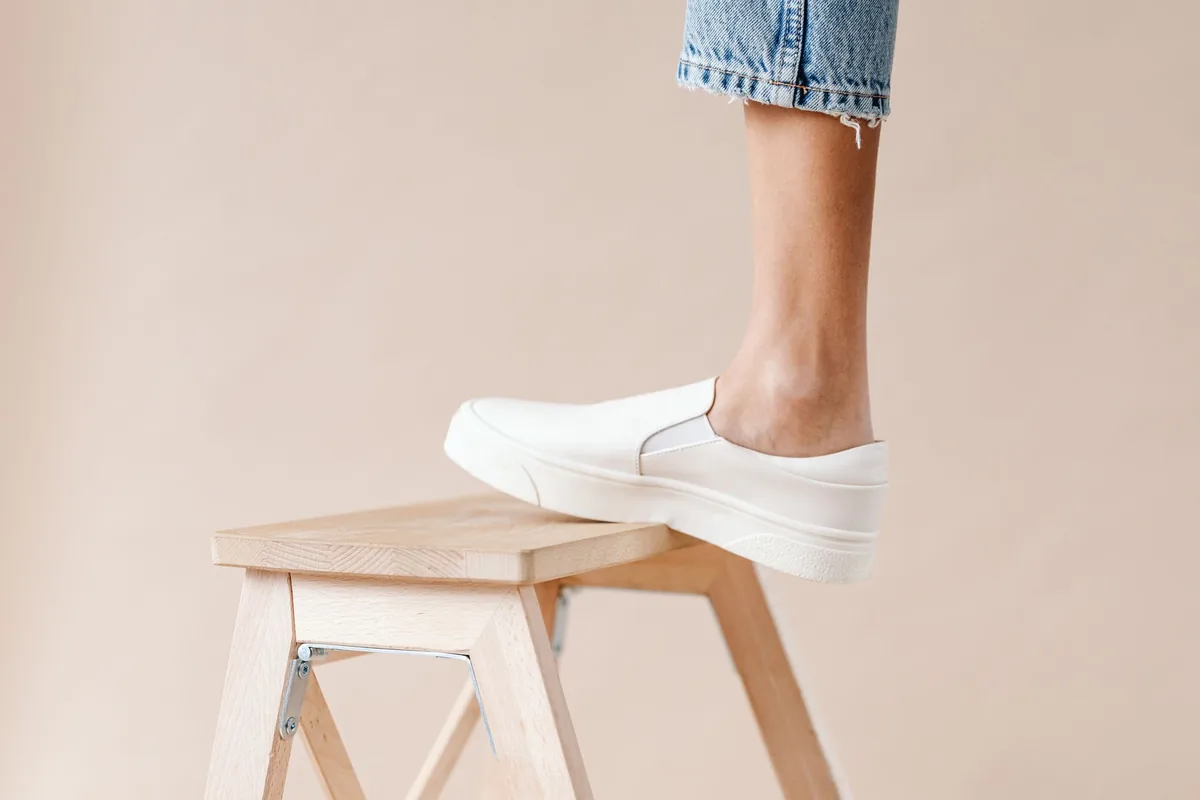 A foot in a white slip-on shoe resting on the top of a plain wooden step stool against a beige wall