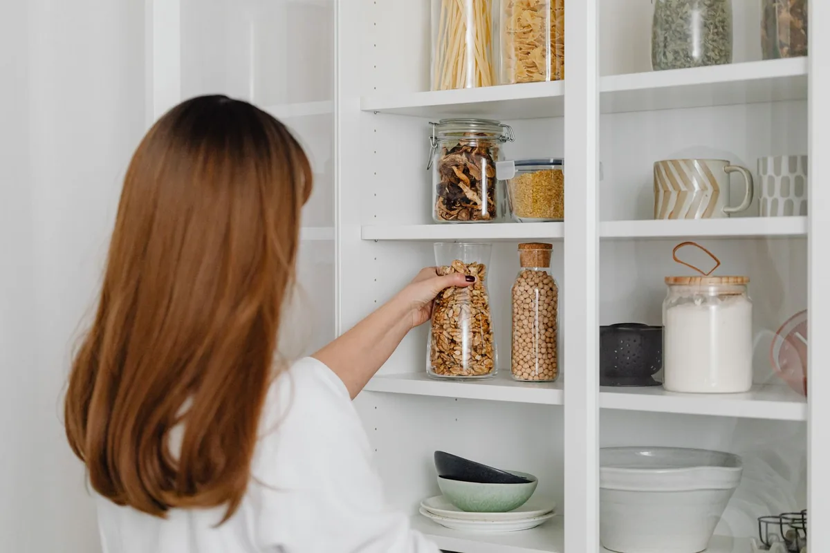A woman with long auburn hair reaching for a glass jar on a white pantry shelf lined with jars, mugs and bowls