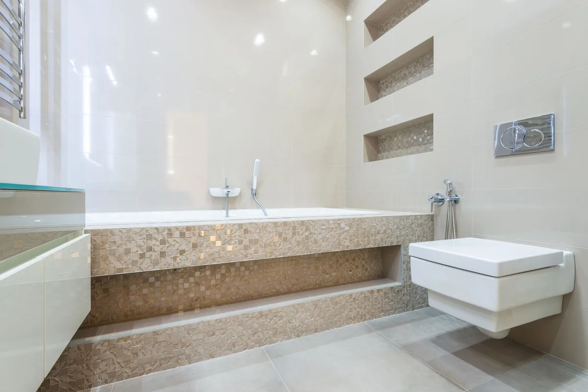 A wall-hung toilet beside a tiled bathtub surround in a bright bathroom with light walls and a chrome flush plate