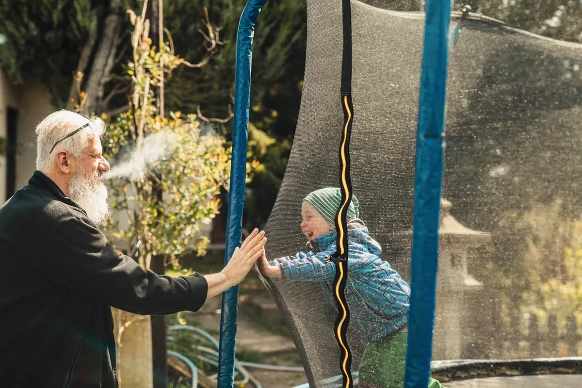 An older man outside a trampoline enclosure pressing his palm to a laughing child's hand through the safety net in a sunny garden