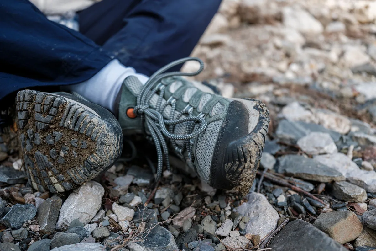 Two mud-speckled low hiking shoes resting on loose gravel, one turned to show the lugged sole