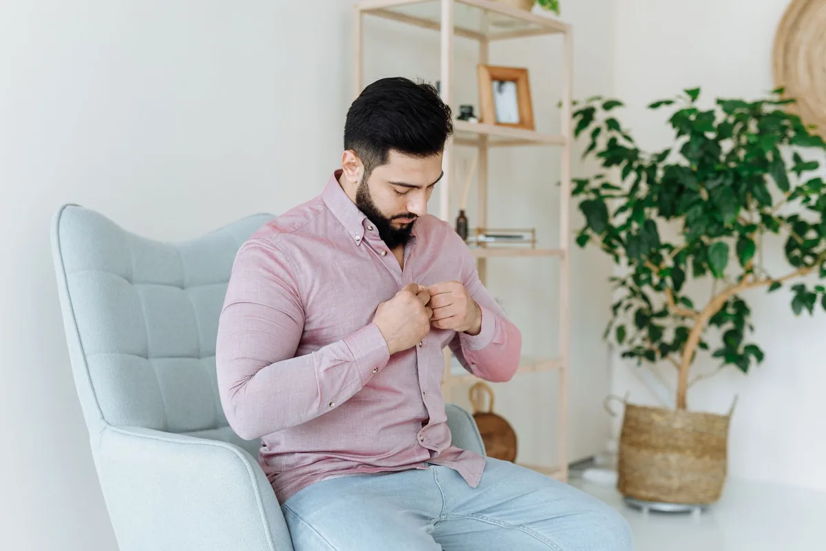 A man buttoning a pink shirt while sitting in an armchair