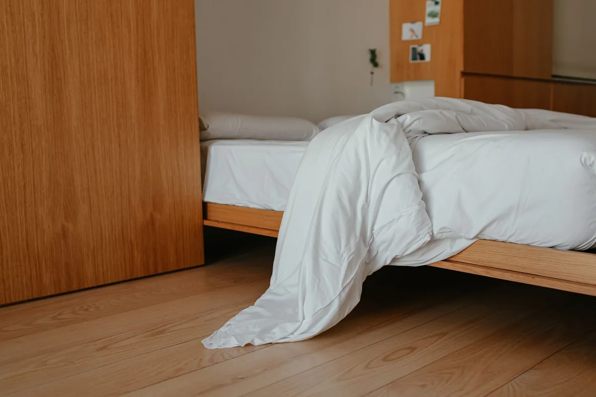 A white duvet slipping off the side of a low wooden bed onto the floor