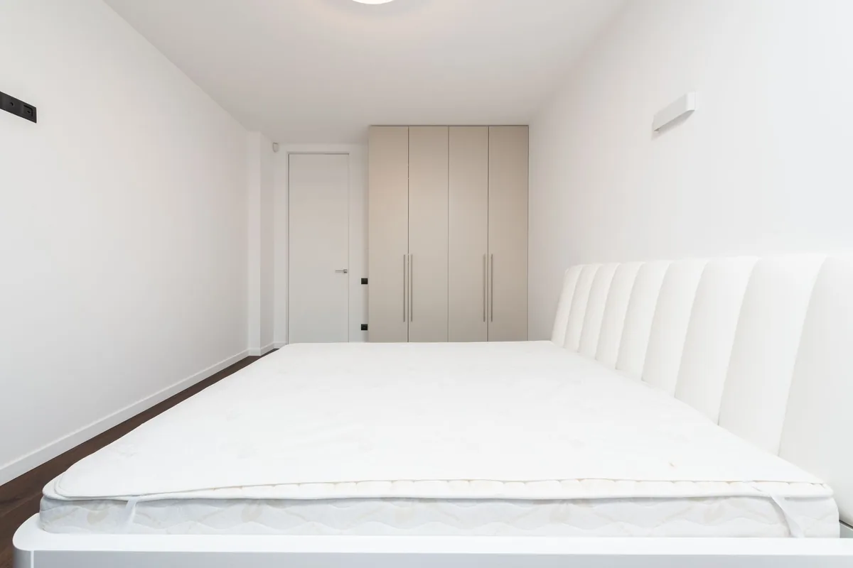 A bare white mattress with a thin topper on a white upholstered bed frame in an empty bedroom