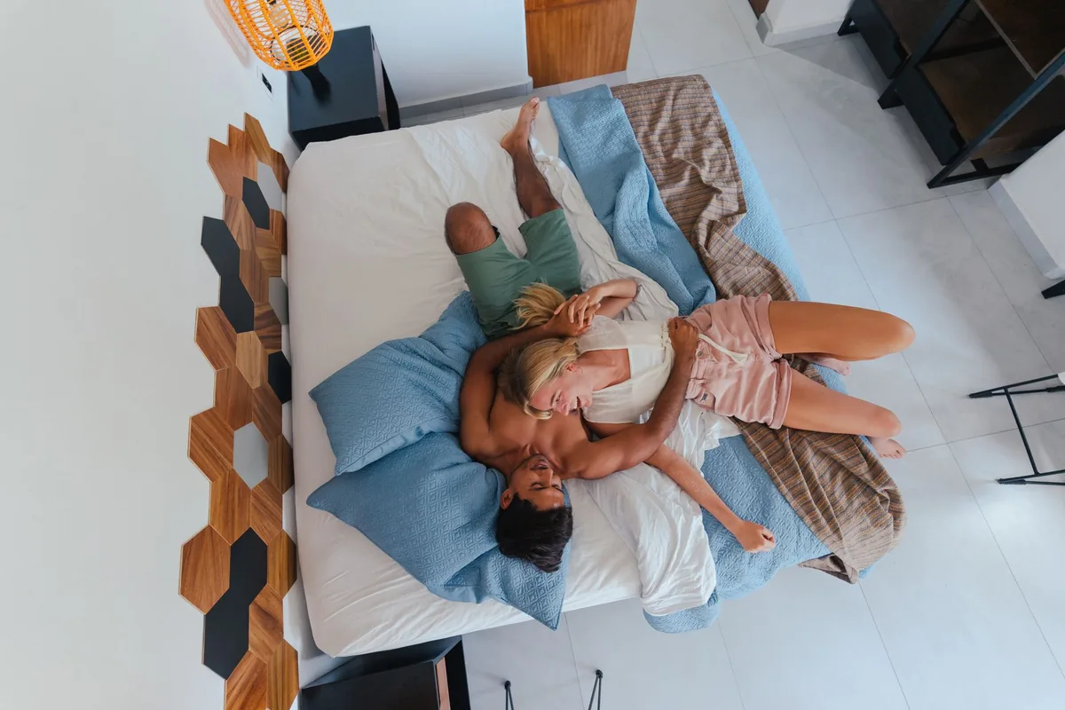 A couple lying together on a large made bed, seen from above, with blue pillows and a striped throw