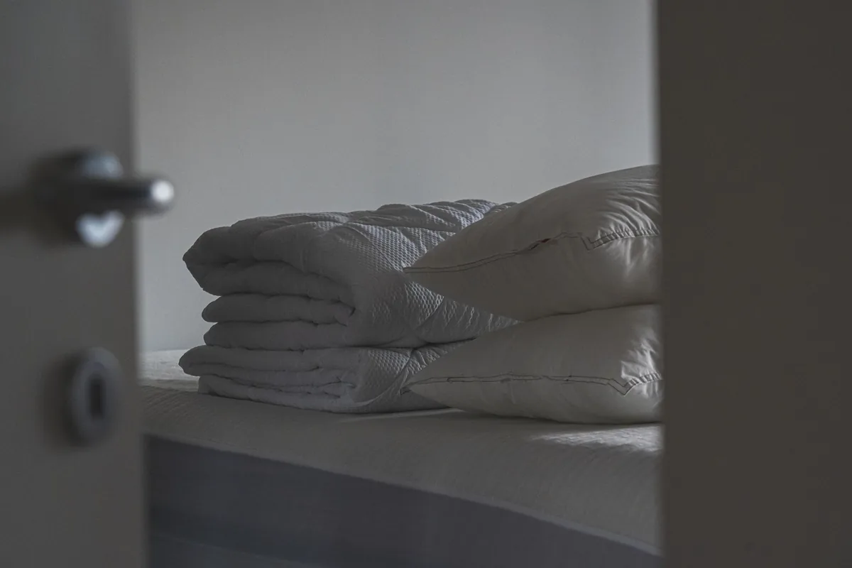 A folded white quilt and two pillows on a bed, seen through a half-open door