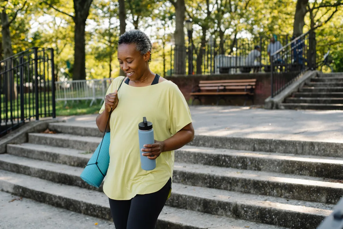 A woman in activewear walking outdoors with a yoga mat and water bottle