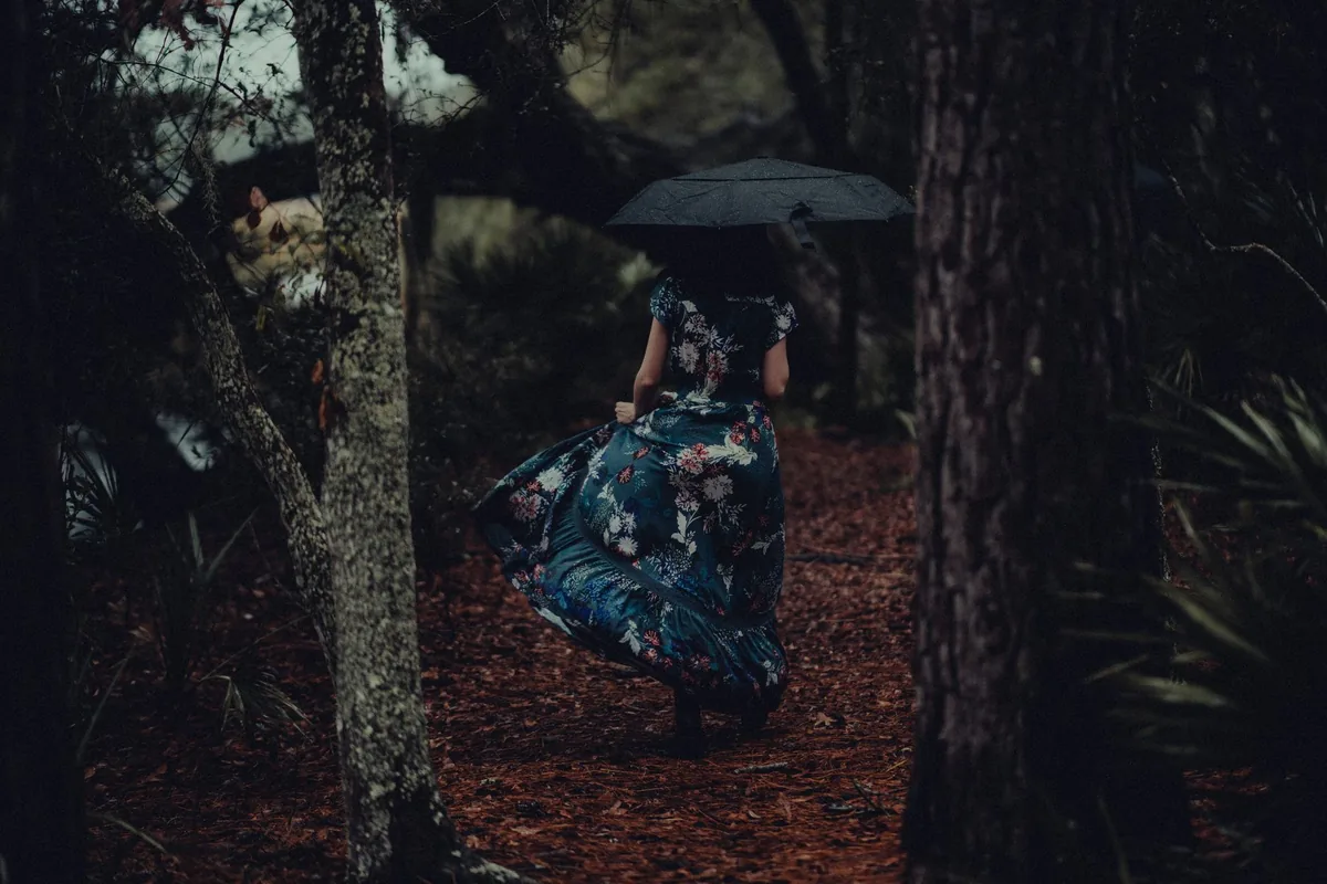 A woman in a long, dark floral dress with short sleeves walking away along a forest path under a black umbrella