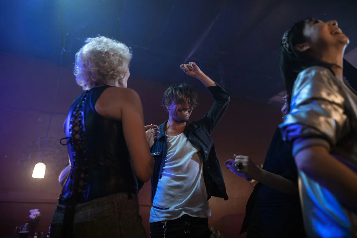 People dancing under blue and purple lights in a dim bar, one in a sleeveless black top with lacing down the back