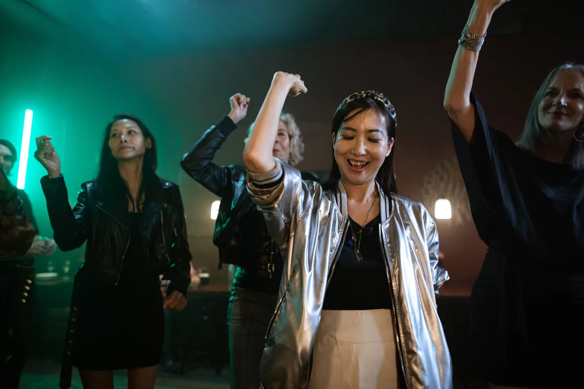 A group of women dancing with their arms raised under green and amber lights, one in a silver jacket over a black top