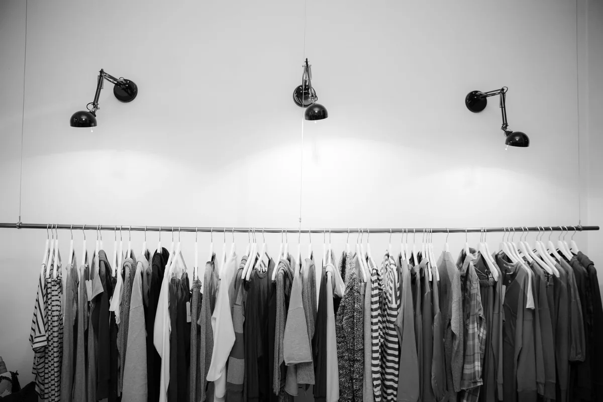 A black and white photo of plain shirts and dresses on white hangers along a clothes rail under three wall lamps