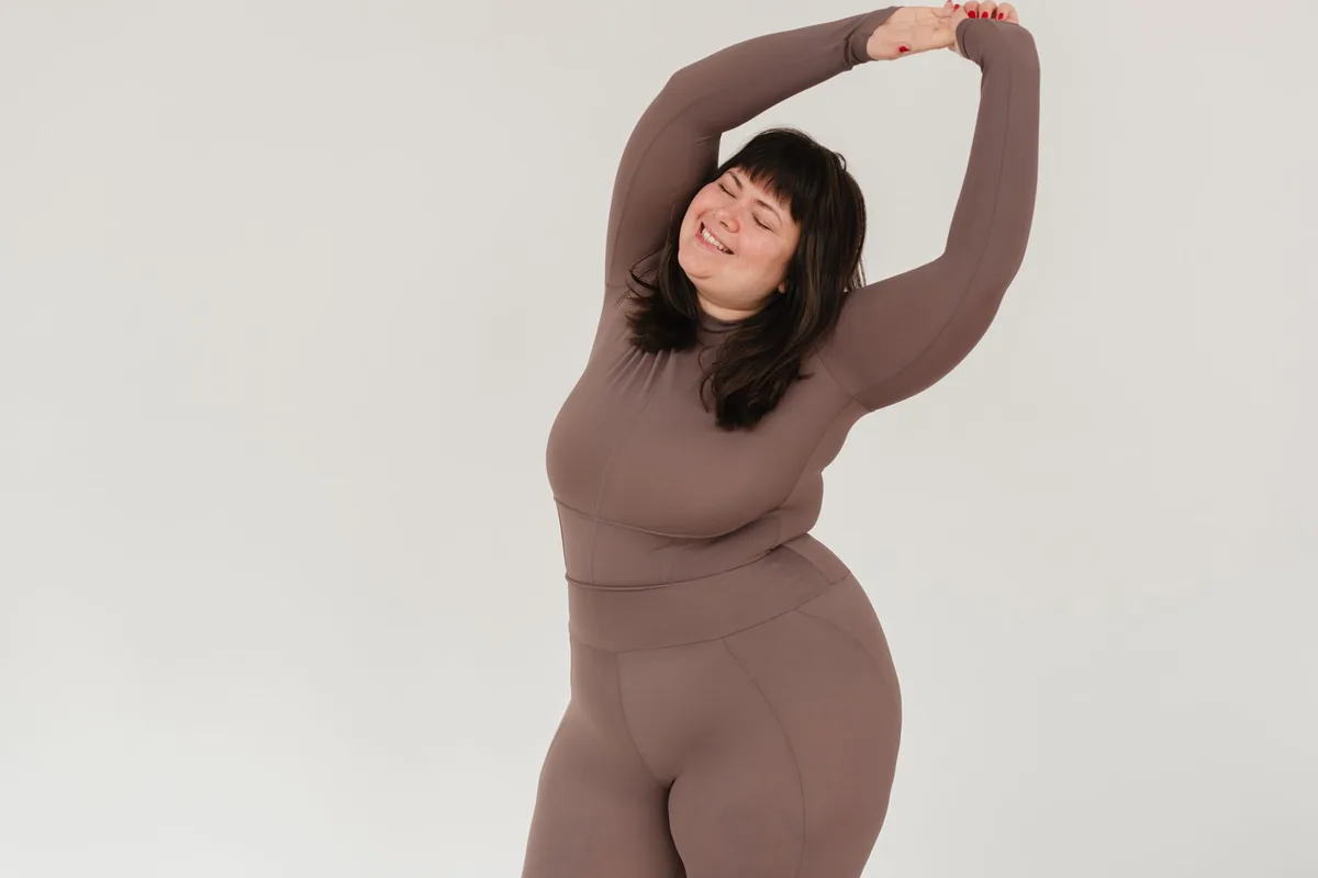 A plus-size woman in brown leggings and a long-sleeve top stretching her arms overhead