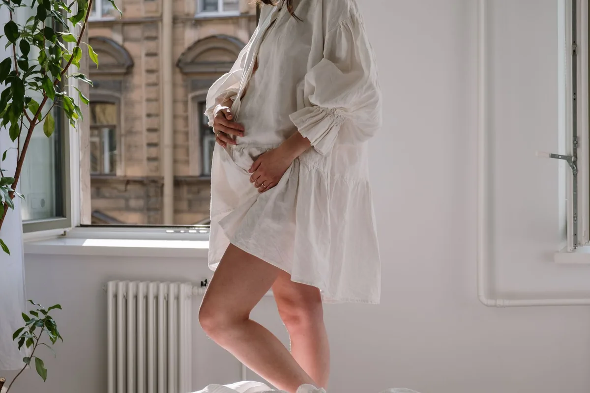 A pregnant woman in a loose white dress standing by an open window
