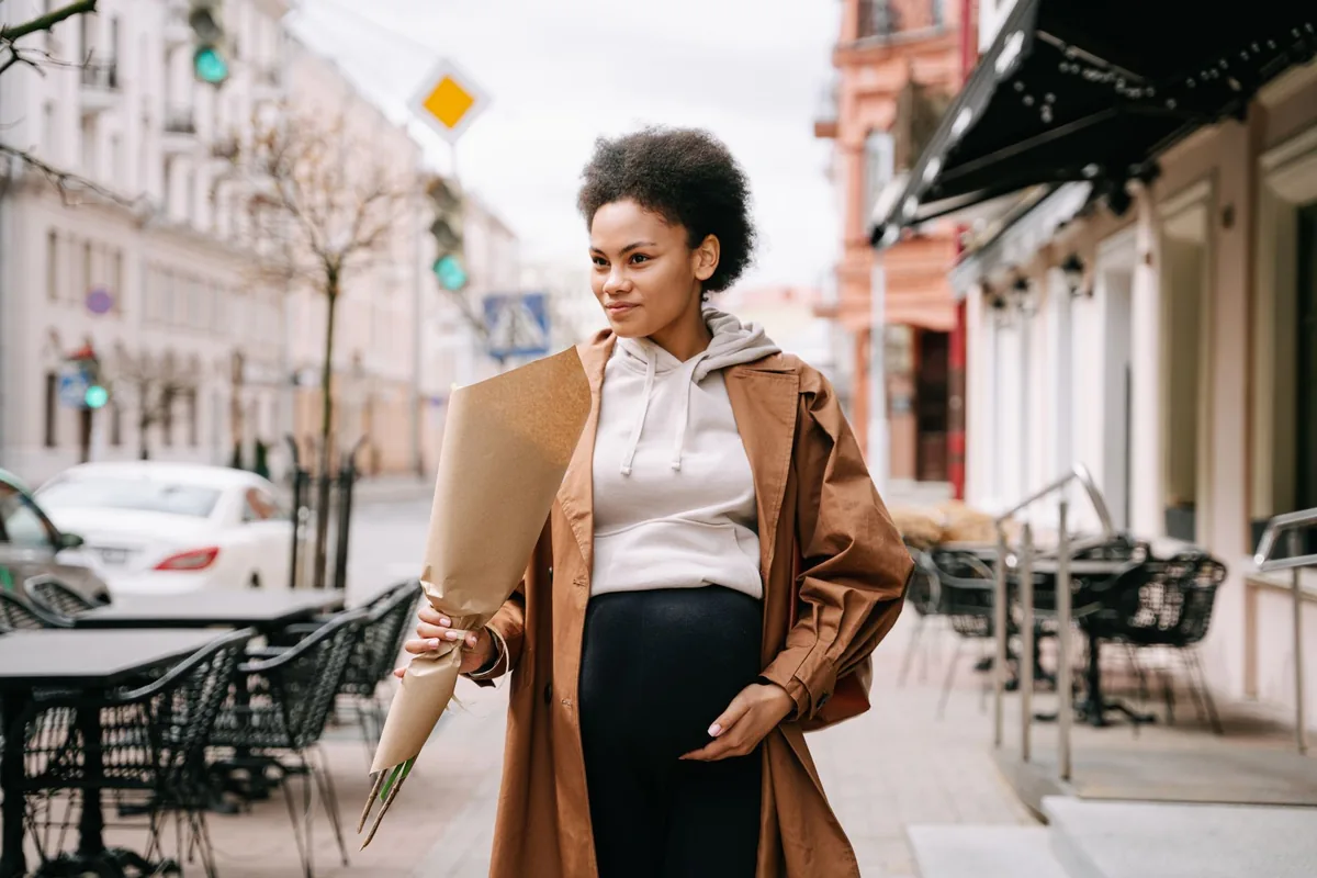 A pregnant woman in a long coat walking down a city street holding flowers
