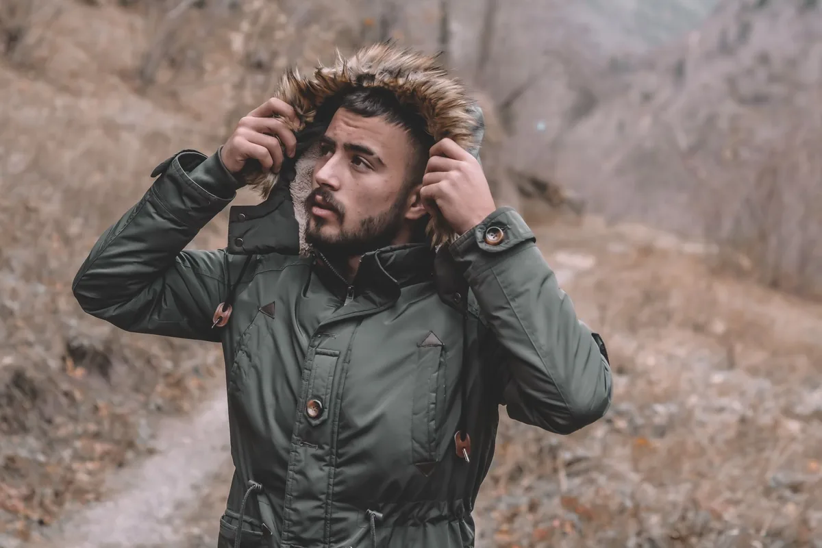A man pulling up the fur-trimmed hood of a green parka on a winter trail