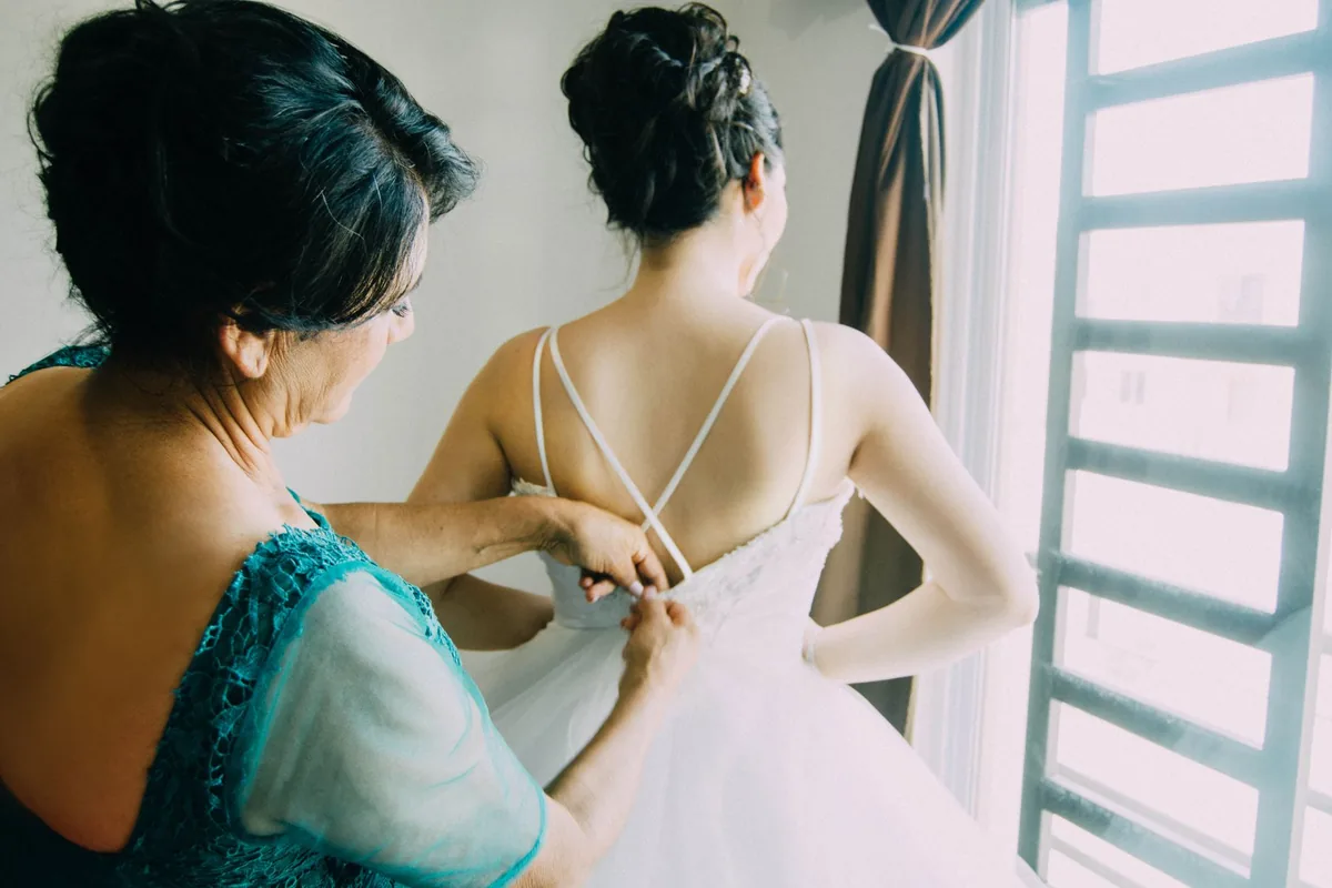 A woman in a teal lace dress fastening the back of a bride's white gown by a sunlit window