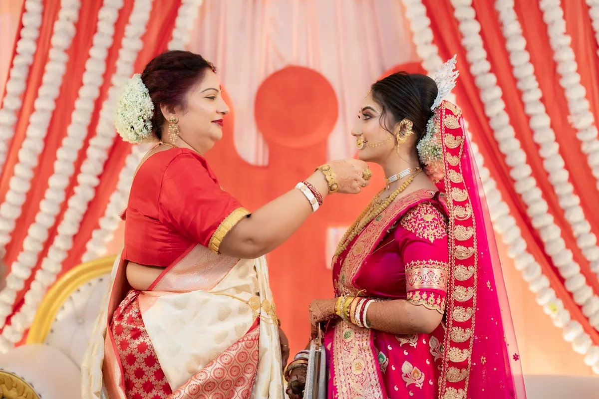 A mother in a red and cream sari touching the face of a bride in pink and red bridal wear at a wedding ceremony