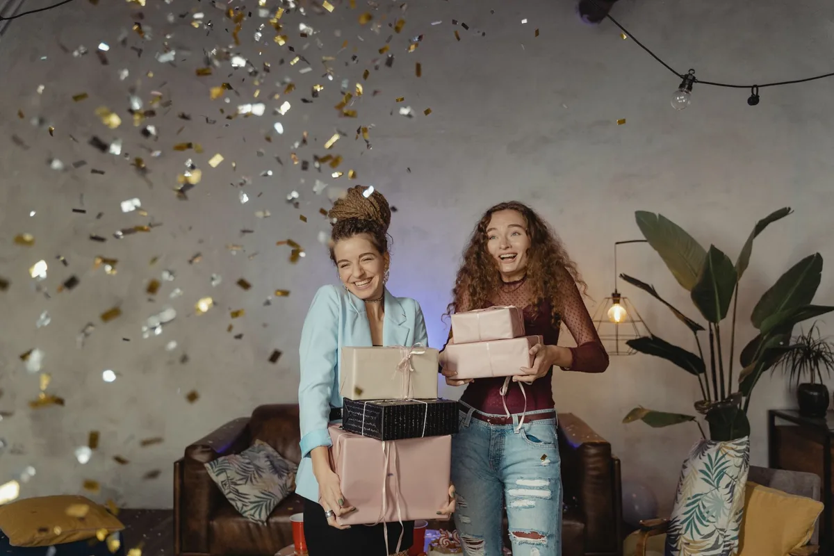 Two women laughing under falling confetti while carrying wrapped gifts