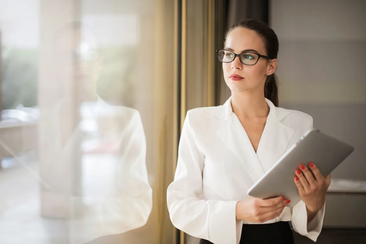 A woman in a dark jacket standing by a window holding a tablet
