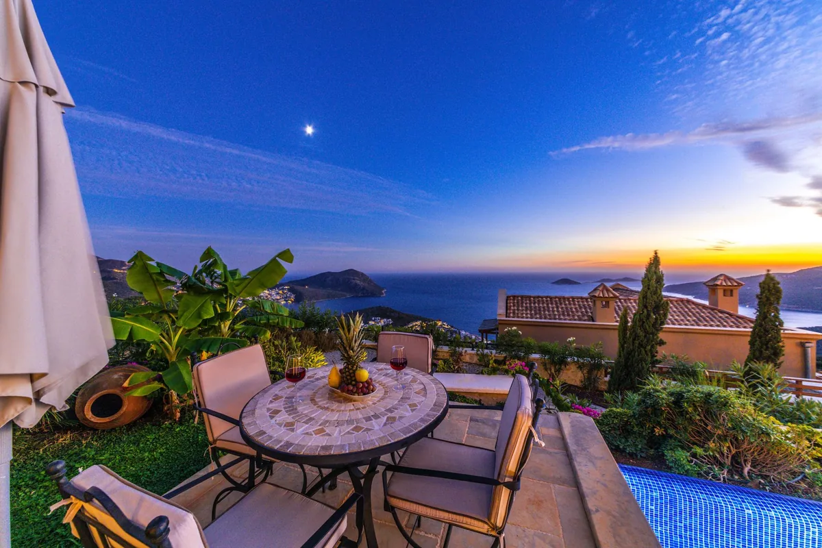 A round mosaic table set with two glasses of red wine on a garden terrace overlooking the sea at dusk