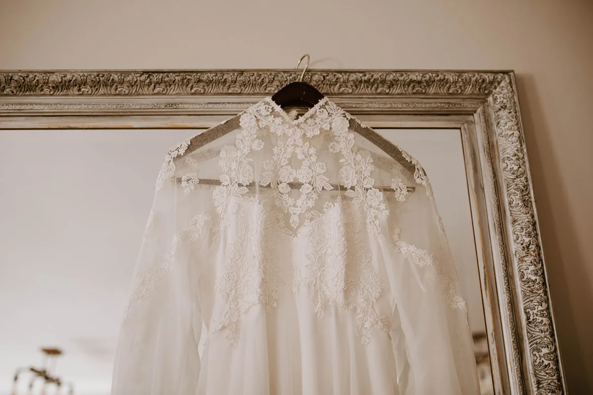 A lace wedding dress with sheer long sleeves hanging from a hanger in front of an ornate gilded mirror