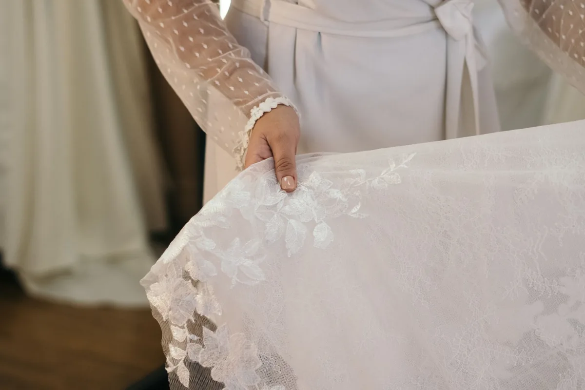 A woman in a sheer dotted long-sleeve top holding up the lace hem of a white wedding gown