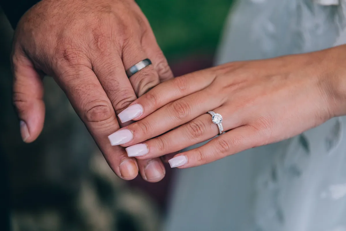 A man's hand with a wide metal band beside a woman's hand with a narrow solitaire ring