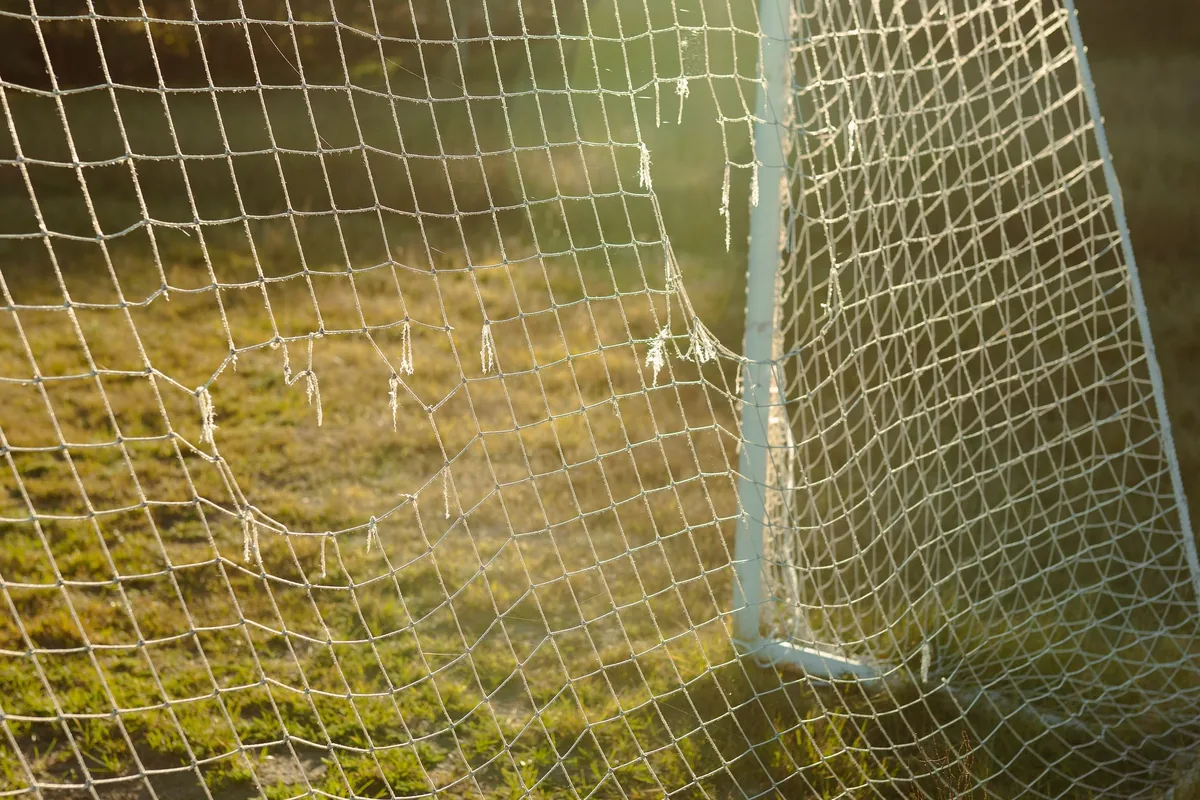 Close-up of a white soccer goal net with worn holes, lit by low sun over a grass field