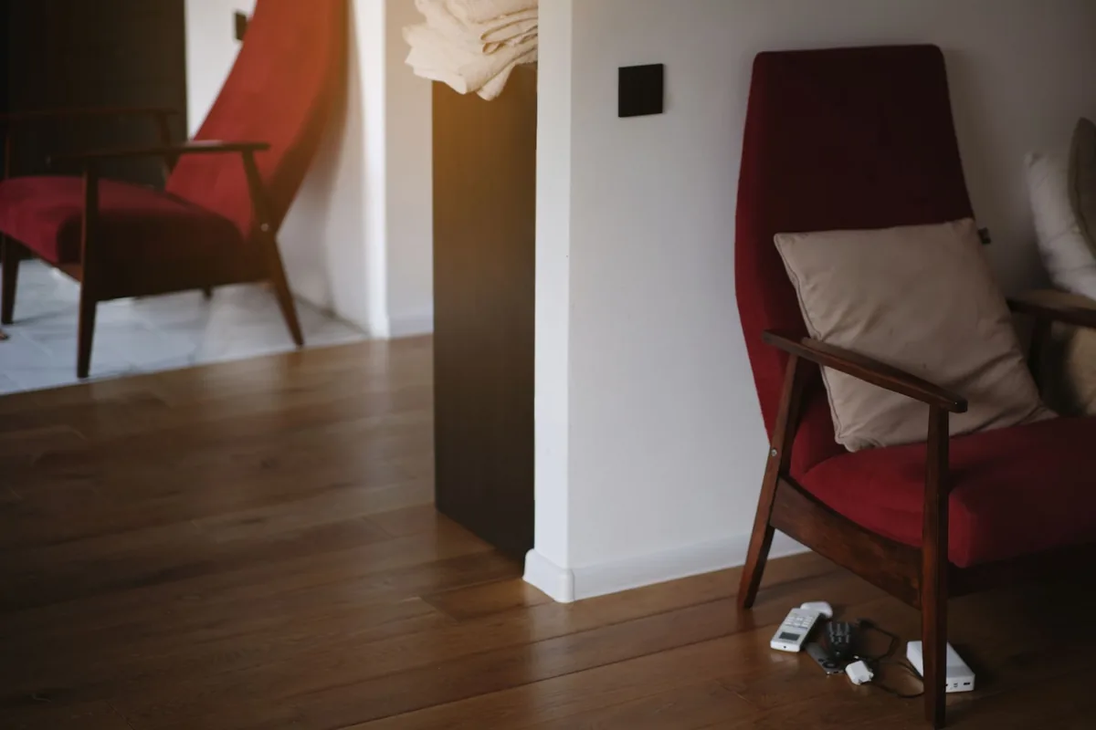 A red upholstered armchair with wooden arms and a beige cushion against a white wall, with a second matching chair in the next room