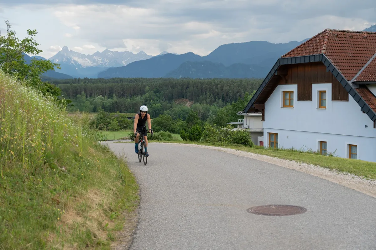 A cyclist in a helmet riding uphill on a narrow paved road past a white house, with forest and mountains behind