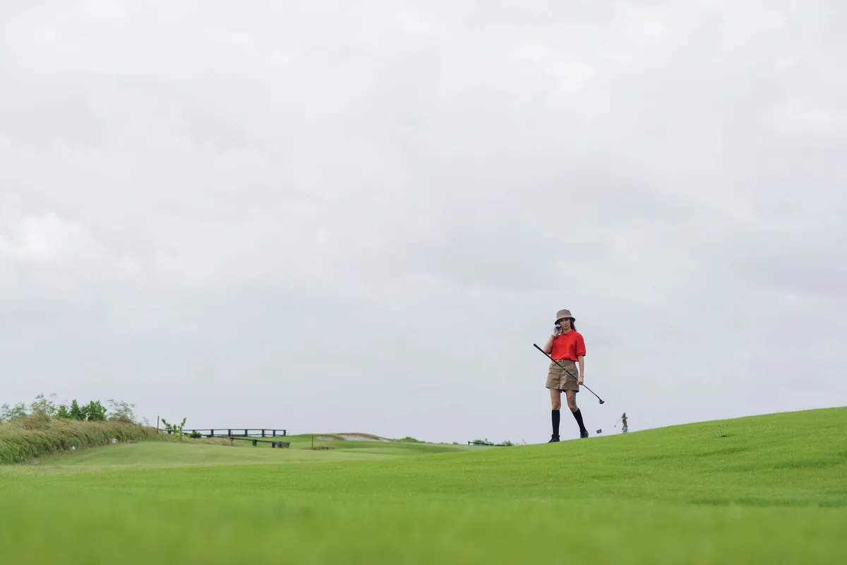 A golfer in a red polo shirt and bucket hat walking across a grassy slope on a golf course, holding a club